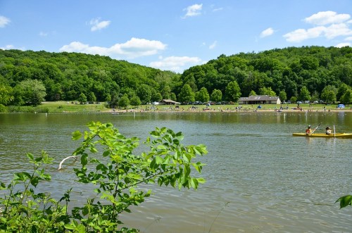 A serene lake surrounded by lush greenery, with people enjoying the beach and kayakers on the water under a blue sky.