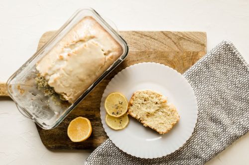 A loaf of lemon cake on a wooden board, with a slice on a plate and lemon halves beside it.
