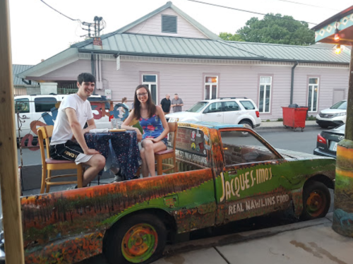 Two people sit at a table on a colorful, painted truck outside a restaurant, with a building and cars in the background.