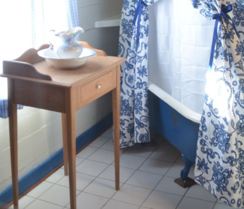 A wooden table with a bowl and pitcher beside a blue and white patterned curtain near a bathtub.