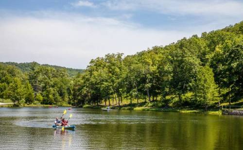 Two people kayak on a calm lake surrounded by lush green trees and hills under a clear blue sky.