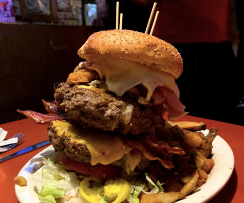 A towering burger with multiple patties, cheese, bacon, and toppings, served with a side of fries on a plate.