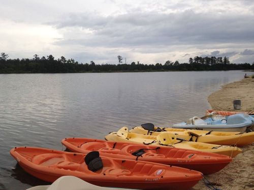 Colorful kayaks lined up on a sandy shore by a calm lake under a cloudy sky.