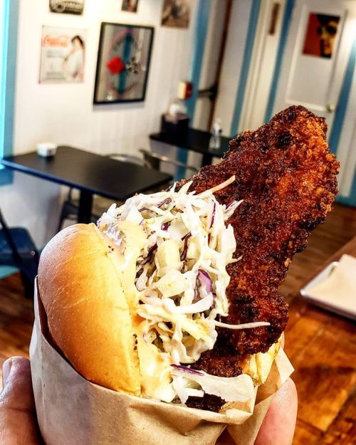 A hand holding a crispy fried chicken sandwich topped with coleslaw, set in a casual dining space.