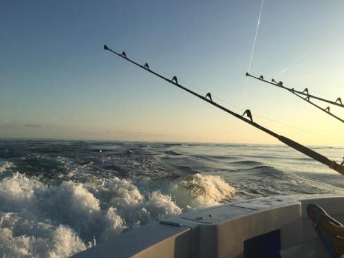Fishing rods extend over the side of a boat, with waves and a serene sunset in the background.