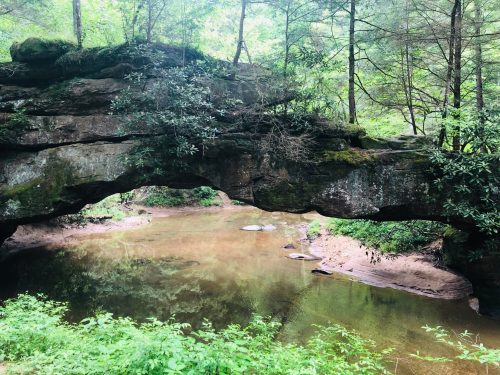 A natural stone arch over a calm river, surrounded by lush greenery and trees.