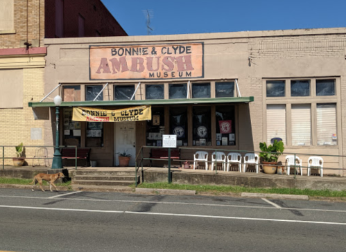 Exterior of the Bonnie & Clyde Ambush Museum, featuring a sign and empty chairs outside, with a dog walking by.