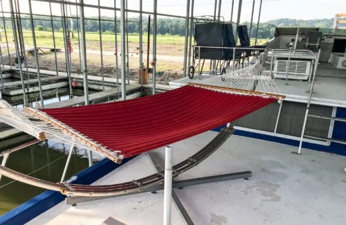 A red hammock is set up on a boat deck, surrounded by a metal frame and overlooking a calm waterway.