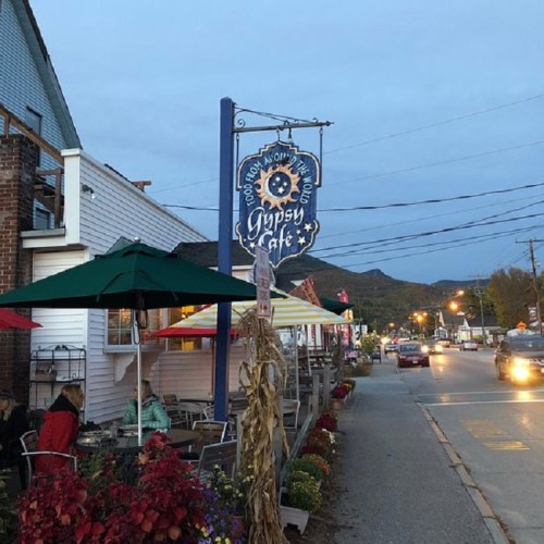 A cozy street view of Gypsy Cafe with outdoor seating, colorful umbrellas, and autumn decorations at dusk.