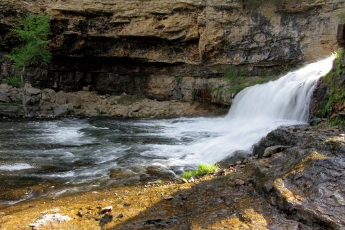 A serene waterfall cascades over rocky terrain into a calm river, surrounded by cliffs and greenery.