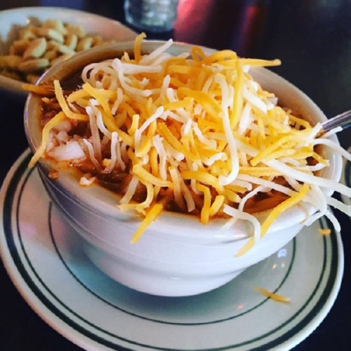 A bowl of chili topped with shredded cheese, served on a plate with a side of peanuts in the background.