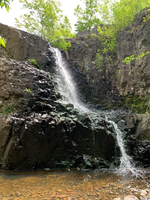 A serene waterfall cascading over dark rocks, surrounded by lush green foliage and a calm pool below.