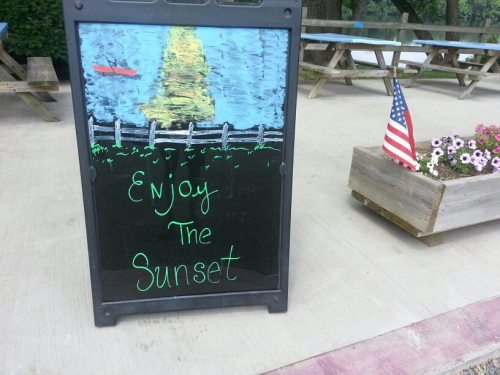 A chalkboard sign by a lake reads "Enjoy The Sunset," with a drawing of a sunset and a small American flag nearby.