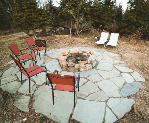A stone fire pit surrounded by red and white chairs in a grassy area with trees in the background.