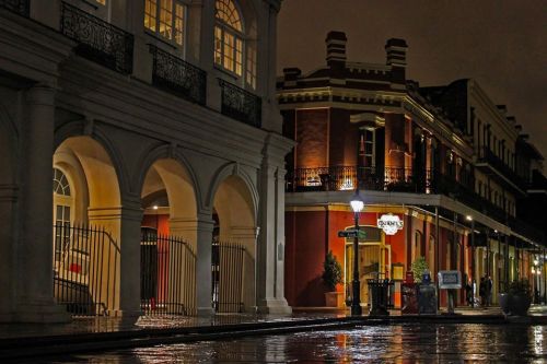 A nighttime street scene featuring historic buildings with arched doorways and warm lights reflecting on wet pavement.