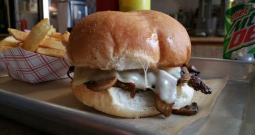 A close-up of a cheesy mushroom sandwich on a bun, served with a side of fries and a soda in the background.