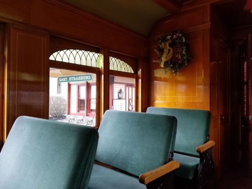 Interior of a vintage train car with green seats and a view of a building labeled "East Strasburg" outside the window.