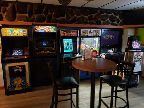 A cozy arcade corner featuring classic games like Pac-Man and Star Wars, with a table and chairs nearby.