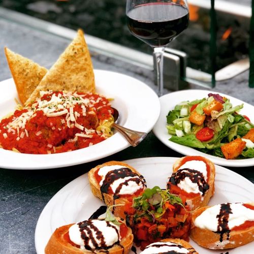 A plate of pasta with marinara sauce and cheese, a side salad, bruschetta drizzled with balsamic, and a glass of red wine.