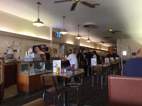 A cozy diner interior with tables, a counter, and patrons enjoying their meals. Ceiling fans and retro decor are visible.
