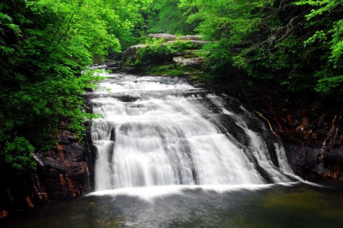 A serene waterfall cascades over rocks, surrounded by lush green trees in a tranquil forest setting.