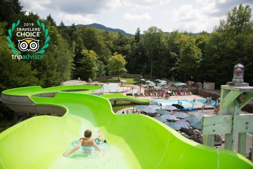 A child slides down a green water slide at a pool area surrounded by trees and lounge chairs.