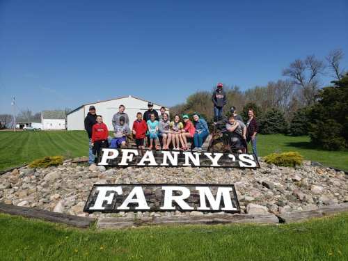 A group of people poses on a sign that reads "Pfanny's Farm" in a sunny outdoor setting with green grass.