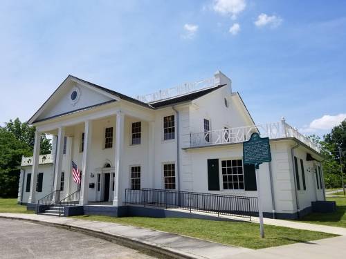 Historic white building with columns, an American flag, and a green historical marker under a blue sky.