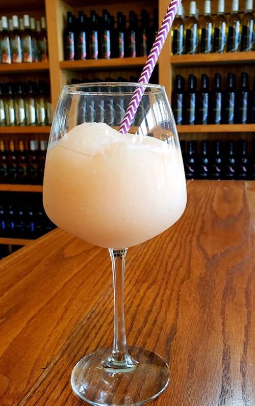 A frosty peach-colored drink in a large glass with a striped straw, set against a backdrop of shelves filled with bottles.