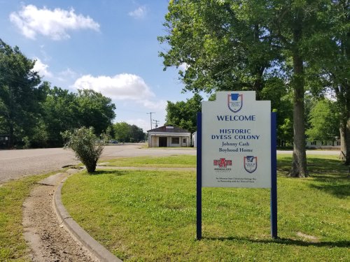 Sign welcoming visitors to the Historic Dyess Colony, Johnny Cash Boyhood Home, surrounded by greenery and a quiet road.