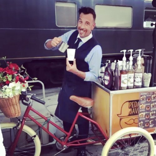A barista in a suit serves coffee from a bicycle cart adorned with flowers, near a train station.