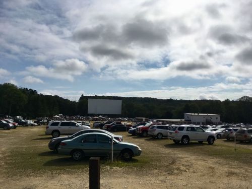 A crowded drive-in movie theater parking lot with a large screen and cloudy sky in the background.