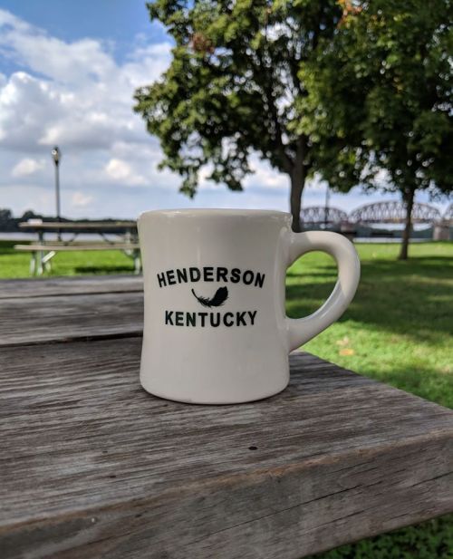 A white mug with "HENDERSON KENTUCKY" printed on it, sitting on a wooden table in a park with trees in the background.