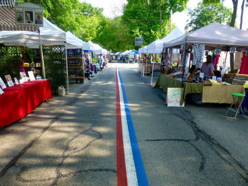 A vibrant street market with colorful vendor tents lining both sides of a tree-lined road marked with red, white, and blue stripes.