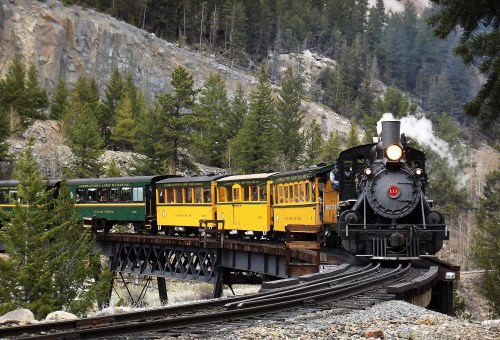 A vintage steam train with yellow and green cars crosses a bridge surrounded by lush trees and rocky terrain.