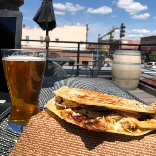 A plate with a grilled quesadilla and a glass of beer on a patio table, with a sunny sky in the background.