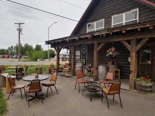 Outdoor seating area with wooden tables and chairs, surrounded by planters and a rustic building.