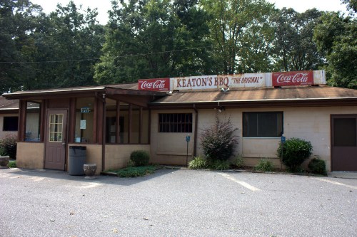 Exterior of Keaton's BBQ restaurant with a Coca-Cola sign, surrounded by trees and a gravel parking area.