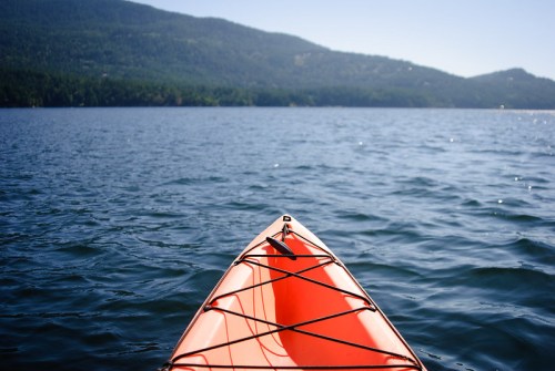 A close-up of an orange kayak's bow on calm water, with a mountainous landscape in the background under a clear sky.