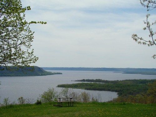 A serene view of a river surrounded by greenery, with a picnic table in the foreground under a cloudy sky.