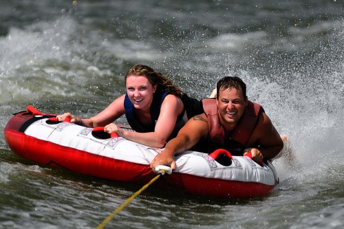 A man and woman enjoy tubing on a lake, laughing as they glide over the water, creating splashes behind them.