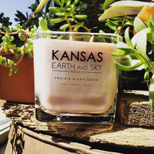 A candle labeled "Kansas Earth and Sky Candle Co." with a prairie wildflower scent, surrounded by green plants.