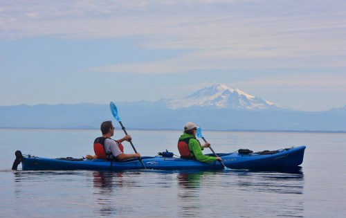 Two kayakers paddle on calm water with a snow-capped mountain in the background under a clear sky.