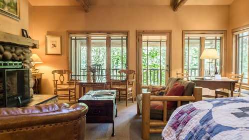 Cozy living room with a stone fireplace, wooden furniture, and large windows overlooking a forested view.