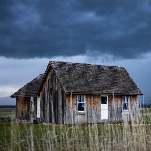 A rustic wooden house stands in a grassy field under a dramatic, cloudy sky.