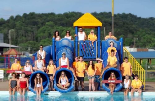 A group of lifeguards in orange shirts poses by a water slide at a pool, with mountains in the background.