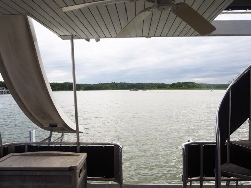 View of a calm lake from a boat, featuring a slide and spiral staircase, with cloudy skies and green hills in the background.