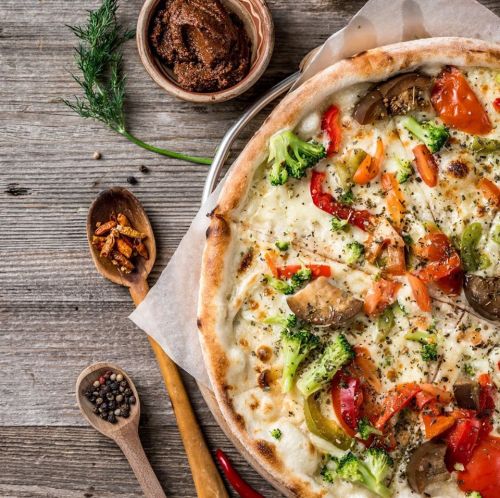A freshly baked vegetable pizza on a wooden table, surrounded by spices and a bowl of sauce.