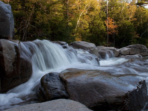 A serene waterfall cascades over smooth rocks, surrounded by lush green trees in a tranquil forest setting.