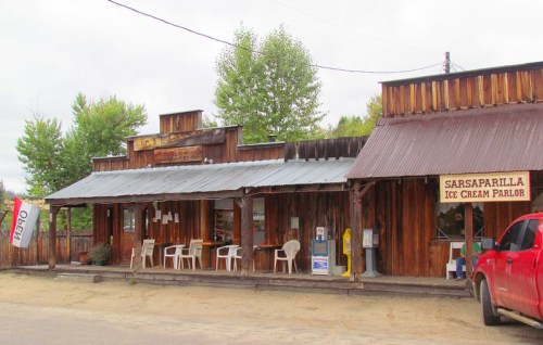 A rustic wooden building with a sign for "Sarsaparilla Ice Cream Parlor" and outdoor seating.